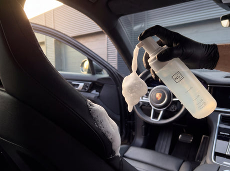 Schwarz behandschuhte Hand pumpt fein-bubbly Schaum mit Koch-Chemie Foamer Bottle auf schwarze Lederlehne im Porsche-Innenraum
